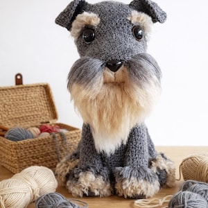 May include: A handcrafted, grey and tan Schnauzer dog plush toy with large black eyes and a fluffy beard. The dog is sitting on a wooden surface, surrounded by yarn and knitting needles. A woven basket filled with yarn is in the background.