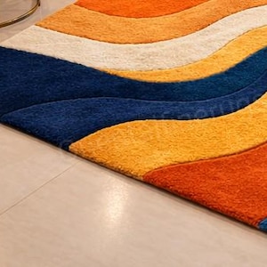 May include: A vibrant area rug with a flowing, wave-like pattern. The rug showcases bands of orange, yellow, white, and navy blue. The rug has a soft, textured feel and is placed on a light-coloured tiled floor.
