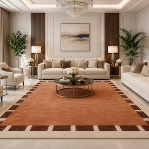 May include: A large rectangular area rug featuring a solid burnt orange centre bordered by a dark brown and cream geometric block pattern. The rug is positioned on a light-coloured floor in a room with cream sofas, armchairs, and a round table.