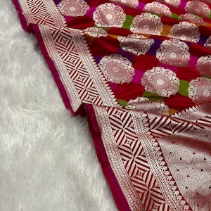 May include: A vibrant textile featuring a multi-colored striped pattern in red, pink, yellow, and green. The fabric is adorned with repeating silver circular floral motifs and a wide, intricate geometric border in silver and deep red.