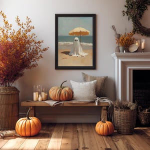May include: A framed wall art print featuring a white ghost standing on a sandy beach under a yellow beach umbrella, with the ocean and sky in the background. The artwork is displayed in a black frame, capturing a surreal scene with soft colors.