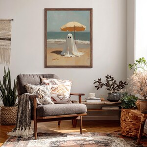 May include: A framed wall art print featuring a white ghost figure standing on a sandy beach under a yellow beach umbrella. The scene depicts the ocean waves in the background with a clear sky. The artwork is enclosed in a dark brown wooden frame.