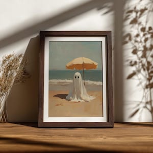 May include: A framed art print featuring a white ghost figure standing on a sandy beach under a yellow beach umbrella. The scene depicts the ocean waves in the background with a clear sky, presented in a dark brown wooden frame on a flat surface.