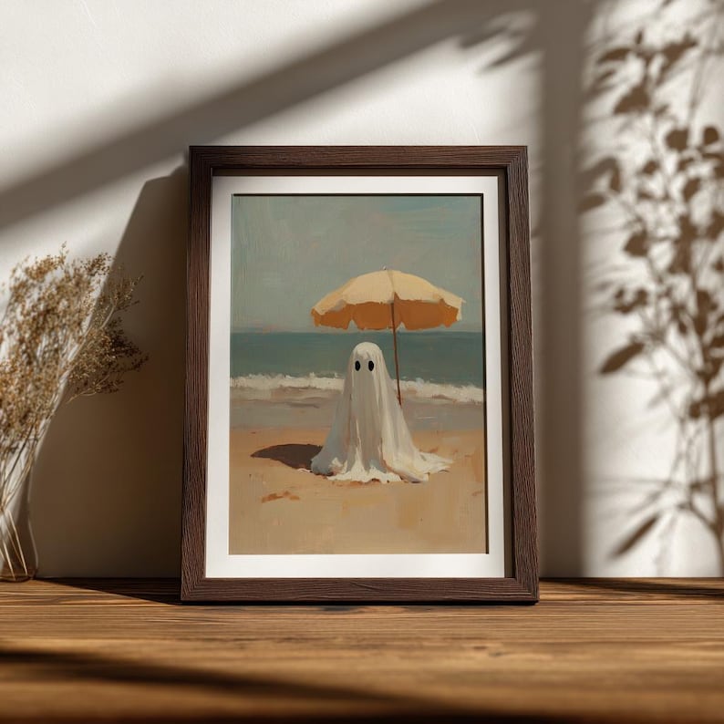 May include: A framed art print featuring a white ghost figure standing on a sandy beach under a yellow beach umbrella. The scene depicts the ocean waves in the background with a clear sky, presented in a dark brown wooden frame on a flat surface.