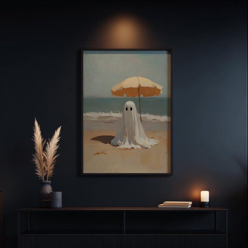 May include: A framed wall art print depicting a white ghost figure standing on a sandy beach under a yellow beach umbrella. The scene features a blue ocean horizon, soft brushstroke textures, and a light-colored sky, presented in a black frame.