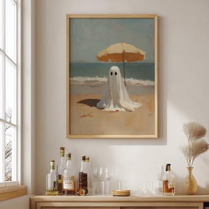May include: A framed wall art print featuring an oil painting of a white ghost figure draped in a sheet, sitting on a sandy beach under a tan beach umbrella. The scene depicts the ocean waves in the background under a soft, muted sky.