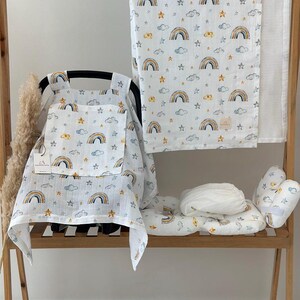 Op de afbeelding: Een babybeddengoedset met een regenboog- en sterrenpatroon op een witte achtergrond. De set bevat een autostoelbekleding, een deken en een kussen. De items worden gepresenteerd op een houten rek. Afmetingen in centimeters.