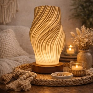 Puede incluir: Una lámpara de cerámica color crema con remolinos y base de madera se encuentra sobre una mesa redonda. La lámpara está iluminada desde dentro, emitiendo un brillo cálido. Una bandeja tejida contiene velas, un cuenco pequeño y cuentas decorativas.