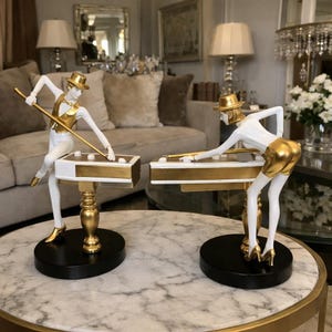 May include: Two decorative figurines of people playing pool. The figurines are white with gold accents, including hats, vests, and shorts. Each figure is posed over a miniature pool table with a black base. The figurines are on a marble-topped table.