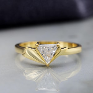 May include: A gold ring featuring a clear, triangular-cut gemstone set in the center. The band has a smooth, polished finish and flares out into a fan-like, pleated design on both sides of the stone, creating a distinct geometric appearance.