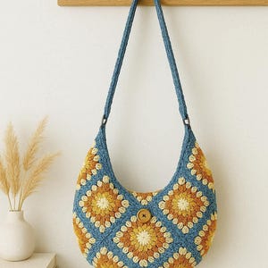 Può includere: Borsa a tracolla all'uncinetto blu e gialla con motivo floreale. La borsa ha una forma arrotondata, una chiusura con bottone in legno e un cinturino blu. La borsa è appesa a un gancio da parete in legno.