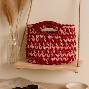 May include: A handmade, two-tone red and pink crochet basket with a handle, resting on a wooden shelf suspended by rope. The basket is a vibrant accent piece. Sunglasses and a small bowl sit on a neutral-colored surface.