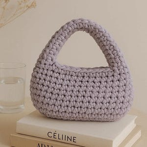 Può includere: Una borsetta all'uncinetto color lavanda, di forma arrotondata e con un manico robusto. La borsa è appoggiata su due libri con i titoli "CÈLIINE" e "AD Victoria". Un bicchiere d'acqua è visibile a sinistra.