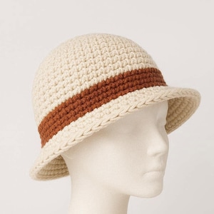 Può includere: Un cappello all'uncinetto color crema con una fascia marrone. Il cappello ha una corona arrotondata e una piccola tesa. Il cappello è esposto su una testa di manichino su sfondo bianco. È un accessorio elegante.