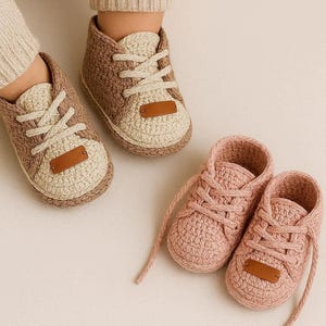 Pode incluir: Tênis de bebê de crochê em duas combinações de cores: bege e marrom, e rosa. Os sapatos têm um design clássico de tênis com cadarços e uma pequena etiqueta marrom na língua. Projetados para bebês.