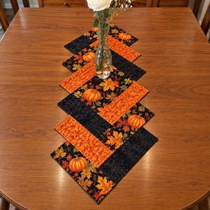 May include: A quilted table runner featuring a geometric pattern of alternating black fabric and orange fabric with pumpkin and autumn leaf prints. The runner is centered on a wooden table with a clear glass vase holding a white flower.