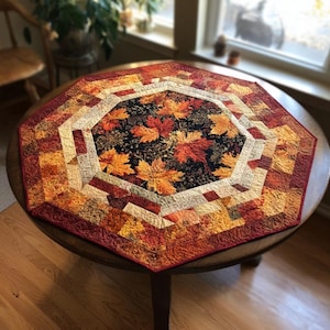 May include: A quilted table topper with a central design of autumn leaves in shades of orange, red, and yellow on a black background. The topper has a geometric pattern with various warm-toned fabrics, creating a cozy, seasonal aesthetic.