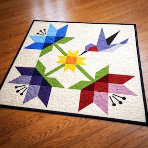 May include: A quilted wall hanging featuring a floral and hummingbird design. The quilt showcases blue, purple, and red flowers with green leaves and a yellow center. A hummingbird is depicted in flight. The quilt has a cream background and a black border.
