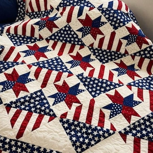 May include: A quilted blanket with a patriotic design. The quilt features alternating patterns of red and white stripes, blue stars, and red and blue star shapes on a cream background. The quilt is likely handmade.