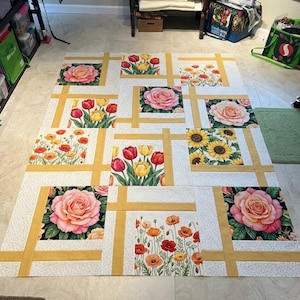 May include: A floral quilt in progress, featuring squares with rose, tulip, poppy, and sunflower designs. The squares are framed with gold-colored fabric and set against a white background with a subtle pattern. The quilt is laid out on a light-colored floor.