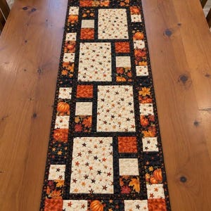 May include: A rectangular quilted table runner featuring a patchwork design of cream fabric with small brown and orange stars, alternating with black fabric squares decorated with orange pumpkins and colorful autumn leaves in shades of red and gold.