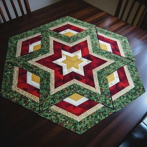 May include: A quilted table runner in a starburst design. The center star is gold, surrounded by red and white star points. The outer border is green with a gold and white trim. The runner is placed on a dark wooden table.