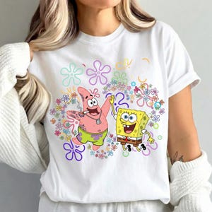 May include: White t-shirt featuring a colorful graphic of SpongeBob SquarePants and Patrick Star. The cartoon characters are surrounded by floral designs in various colors. The shirt has short sleeves and a crew neck.