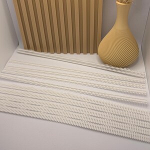 Könnte beinhalten: Eine Ansammlung weißer Kunststoff-Kleidungsanhänger ist auf einer weißen Oberfläche angeordnet. Eine beige Vase mit strukturiertem Design und eine beige Holzplatte befinden sich im Hintergrund.