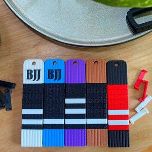 Puede incluir: Cinco llaveros rectangulares de cinturón de BJJ en blanco, azul, morado, marrón y negro, cada uno con un orificio en la parte superior. Los cinturones blanco y azul muestran el texto BJJ. Clips de plástico negro, rojo y blanco están dispersos.