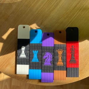 Puede incluir: Cinco marcapáginas rectangulares con iconos de piezas de ajedrez. Cada uno tiene una sección central negra con una pieza de color y bordes a juego en blanco, azul, morado, naranja y rojo. Cada marcapáginas tiene un orificio en la parte superior.