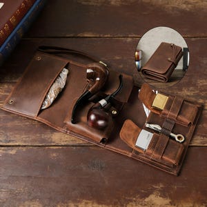 May include: A brown leather tobacco pipe case, open to display two pipes, a lighter, and other accessories. The case is on a wooden surface. An inset shows a closed matching leather wallet.