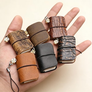 May include: A collection of miniature leather-bound notebooks in various shades of brown and black. Each notebook is secured with a black elastic band and features a decorative charm. The notebooks have different textures, including embossed and crocodile-like patterns.