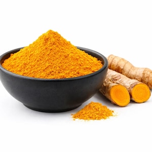 Puede incluir: Un cuenco negro lleno de polvo de cúrcuma amarillo brillante, con un pequeño montón de polvo al lado. Raíces enteras de cúrcuma y una raíz cortada por la mitad están a la derecha, todo sobre un fondo blanco.