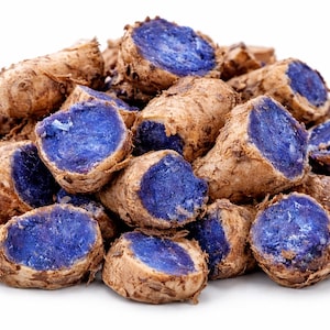 Puede incluir: Un montón de patatas azules frescas y crudas, cortadas para revelar su vibrante pulpa púrpura. Las patatas tienen un exterior marrón rugoso y están dispuestas sobre un fondo blanco. La imagen destaca el contraste de colores.