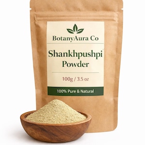 May include: A brown paper bag labelled "BotanyAura Co Shankhpushpi Powder" with a wooden bowl filled with light green powder. The bag states the contents are 100g and 100% Pure & Natural.