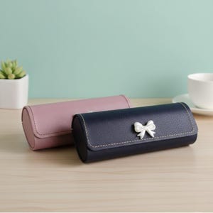 Könnte beinhalten: Zwei zylindrische Etuis, eines rosa und eines marineblau, liegen nebeneinander. Das marineblaue Etui hat eine weiße Schleife. Beide Etuis haben eine genähte Zierleiste. Die Etuis liegen auf einer hellen Holzoberfläche.