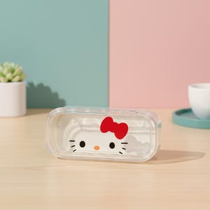 Può includere: Contenitore rettangolare trasparente con un design di Hello Kitty. Il contenitore ha un fiocco rosso e un viso bianco con occhi e baffi neri. Il contenitore è su una superficie di legno.