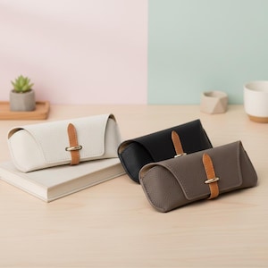 Könnte beinhalten: Drei Leder-Brillenetuis in Creme, Schwarz und Taupe. Jedes Etui hat einen kontrastierenden Riemen in Hellbraun mit einer goldfarbenen Schnalle. Die Etuis werden auf einer hellen Holzoberfläche präsentiert, mit einem Buch und einer kleinen Pflanze im Hintergrund.