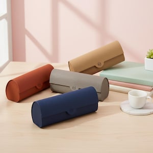 May include: Five cylindrical eyeglass cases in shades of rust, tan, grey, and navy blue. Each case features a rounded top and a snap closure. The cases are displayed on a light wood surface.