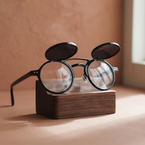 May include: Black round-frame glasses featuring a flip-up mechanism with dark tinted lenses. The eyewear is positioned on a rectangular wooden stand, showcasing clear optical lenses beneath the secondary flip-up sun lenses and thin temples.