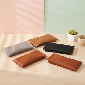 May include: A collection of rectangular eyeglass cases in various colors, including gray, tan, brown, and black. Each case has a textured surface and a silver-colored accent at the opening. The cases are arranged on a light-colored wooden surface.