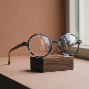Könnte beinhalten: Runde Brille mit einem transparenten grauen Acetatrahmen in polierter Ausführung und hellbraunen Holzbügeln. Die Brille hat klare Gläser und ruht auf einem rechteckigen Block aus dunkelbraunem Holz mit einer sichtbaren Maserung.