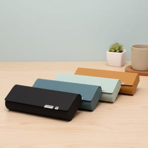 Può includere: Quattro astucci rettangolari con coperchi curvi e chiusure argentate, in fila. Gli astucci sono neri, blu, verde chiaro e marrone chiaro. Sono disposti su una superficie di legno marrone chiaro.