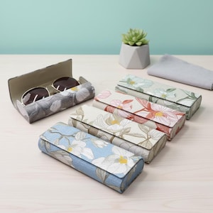 Könnte beinhalten: Eine Sammlung von Brillenetuis mit floralem Muster in verschiedenen Farben, darunter Blau, Grau und Creme. Ein Etui ist geöffnet und zeigt eine Sonnenbrille. Die Etuis sind rechteckig und haben einen Klappverschluss. Im Hintergrund befindet sich eine kleine Sukkulentenpflanze in einem geometrischen Topf.