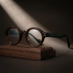 Può includere: Occhiali rotondi con montatura tartarugata marrone scuro e lenti trasparenti. Le aste sono di un colore verde scuro uniforme, con due piccoli rivetti argentati su ciascun lato della montatura frontale. Gli occhiali poggiano su un blocco di legno.