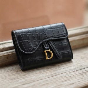 Puede incluir: Una cartera negra con textura de piel de cocodrilo, que presenta un cierre de solapa curvado y un llamativo dije dorado con la letra D sujeto a la correa frontal. El accesorio tiene una forma rectangular estructurada con costuras visibles.