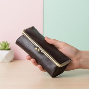 Puede incluir: Un estuche de gafas de cuero marrón oscuro con un cierre dorado. El estuche es rectangular y tiene una superficie texturizada. El cierre es un diseño con bisagras con dos pequeños acentos redondos.