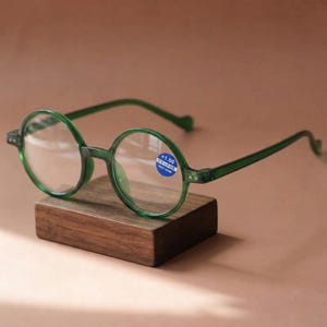 Puede incluir: Gafas de lectura redondas con montura de plástico verde esmeralda translúcido. Las lentes son transparentes y tienen una pequeña pegatina circular azul en la lente derecha con el texto blanco +1.00. Tienen patillas finas y puente clásico.