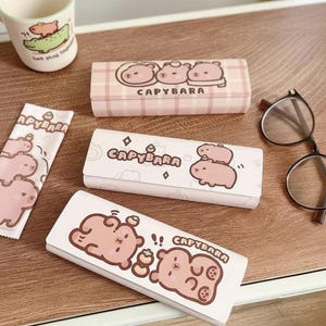 Könnte beinhalten: Drei rechteckige Brillenetuis und ein passendes Reinigungstuch mit Cartoon-Capybara-Illustrationen und dem Text "CAPYBARA". Die Etuis sind hellrosa und weiß und zeigen verschiedene Capybara-Motive, darunter gestapelte und schlafende Figuren.