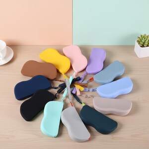 Könnte beinhalten: Eine Sammlung farbenfroher, tropfenförmiger Brillenetuis in verschiedenen Farben, darunter Gelb, Rosa und Blau. Jedes Etui hat eine passende Handschlaufe. Die Etuis sind kreisförmig auf einer hellen Holzoberfläche angeordnet.